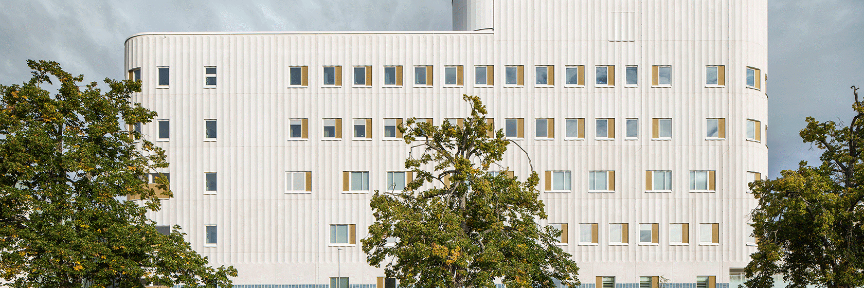 Vit fasad på modern sjukhusbyggnad med grönskande träd i förgrunden