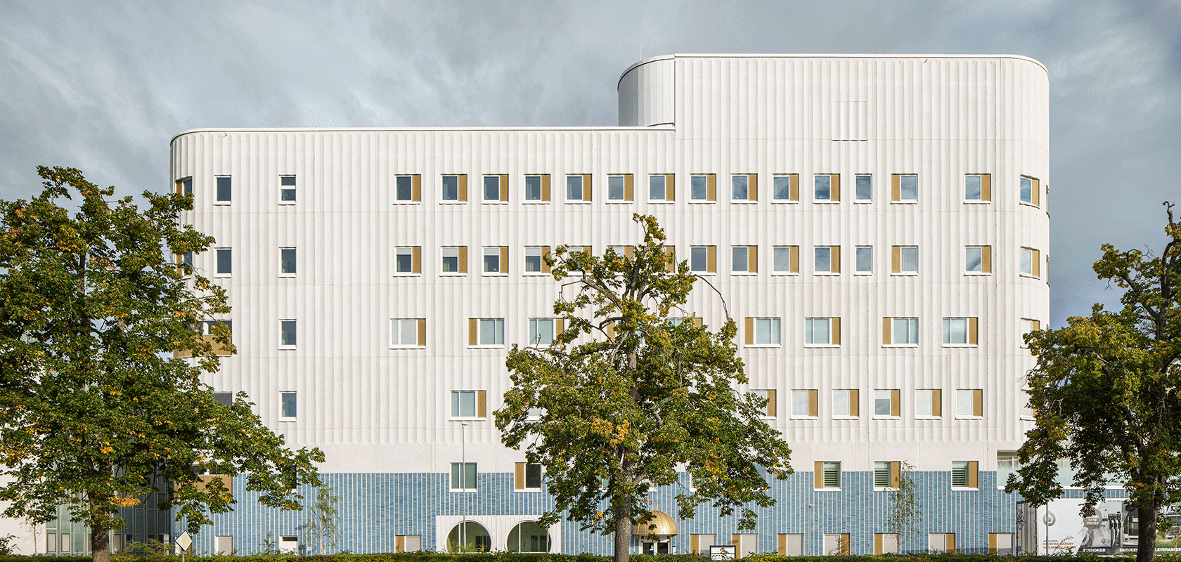 Vit fasad på modern sjukhusbyggnad med grönskande träd i förgrunden