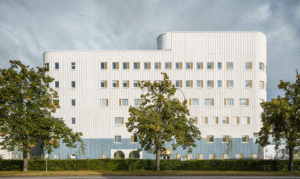 Långsidan på Västerviks nya psykiatrisjukhus. Vit fasad med grönskande träd i förgrunden. Gråblå himmel.