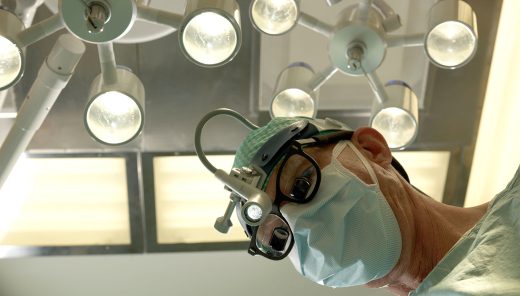 Närbild på kirurg med munskydd och strålkastare ovanför huvudet.