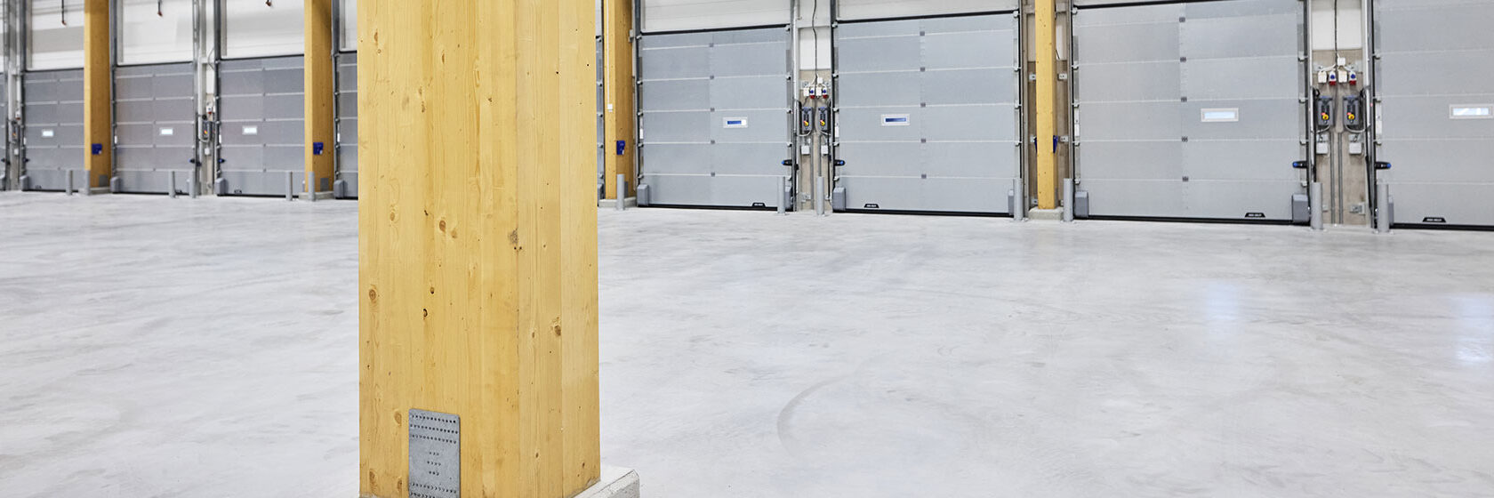 Interiör av logistikbyggnad med silvergrå garageportar i bakgrunden och träbalk i förgrunden