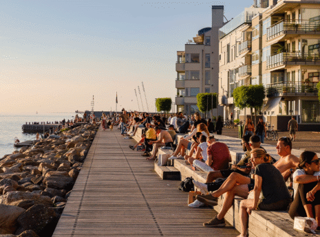 Strandpromenad i stadsmiljö full med människor på träbänkar och flerbostadshus i bakgrunden.
