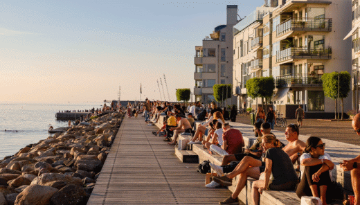Strandpromenad i stadsmiljö full med människor på träbänkar och flerbostadshus i bakgrunden.