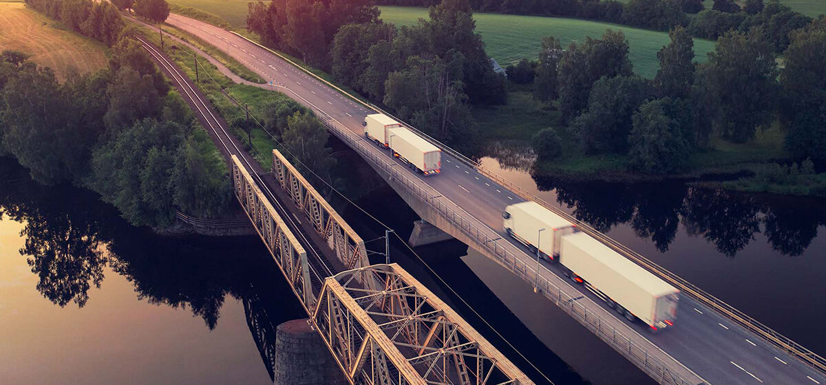 Lastbilar kör över en bro vid solnedgång, med grönskande landskap och järnväg bredvid.