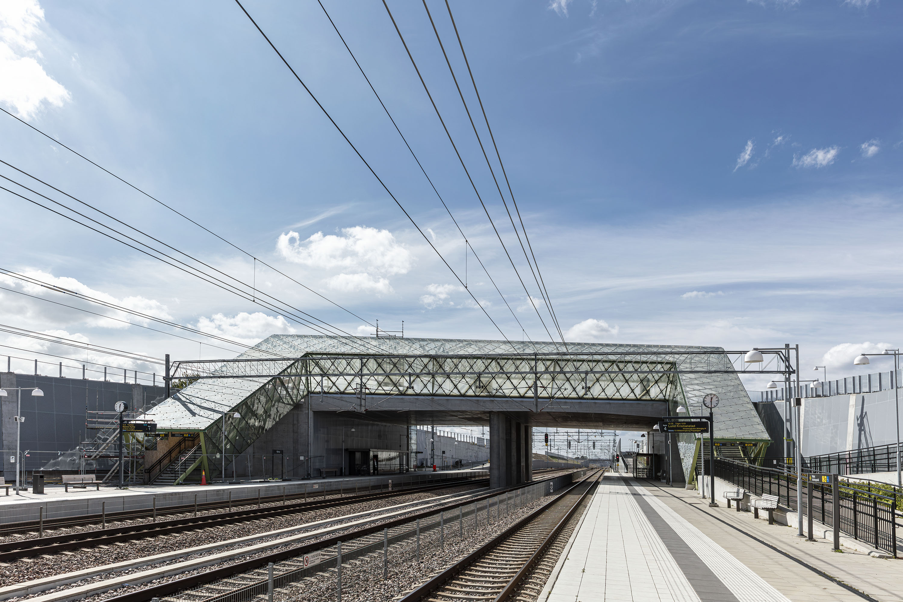 Modern järnvägsstation med gångbro över spåren under en klarblå himmel.
