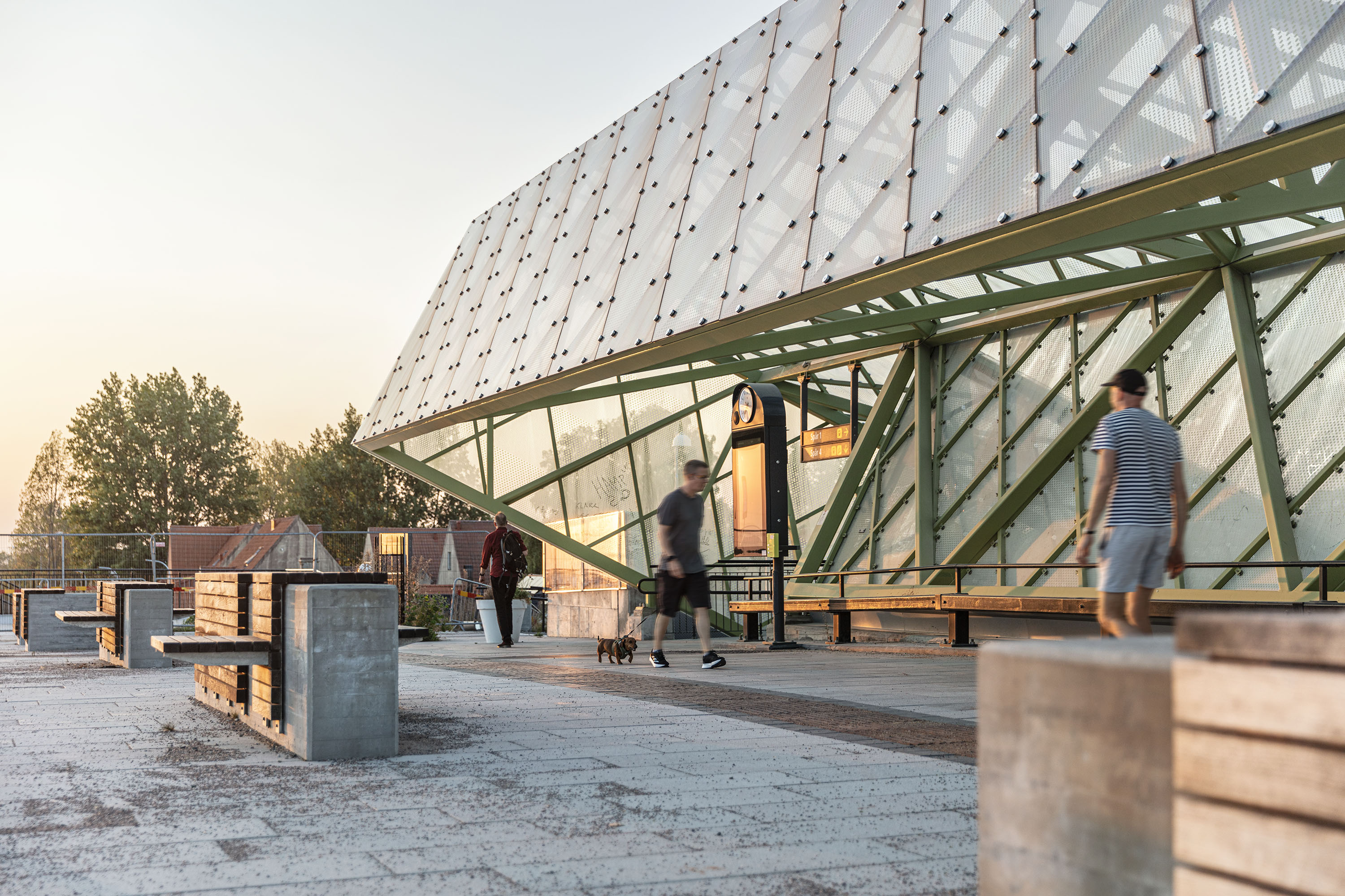 Modern arkitektur med promenadstråk och bänkar, träd och hus i bakgrunden.