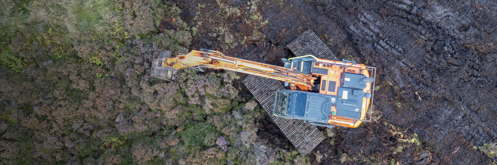 Grävmaskin som arbetar på en byggarbetsplats, omgiven av skog och jord, sedd från ovan.