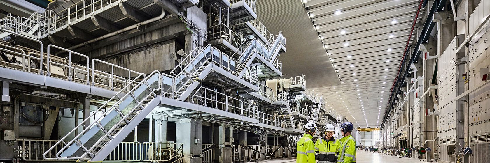 Tre arbetare i skyddsutrustning diskuterar i en modern fabriksmiljö med stora industriella maskiner.