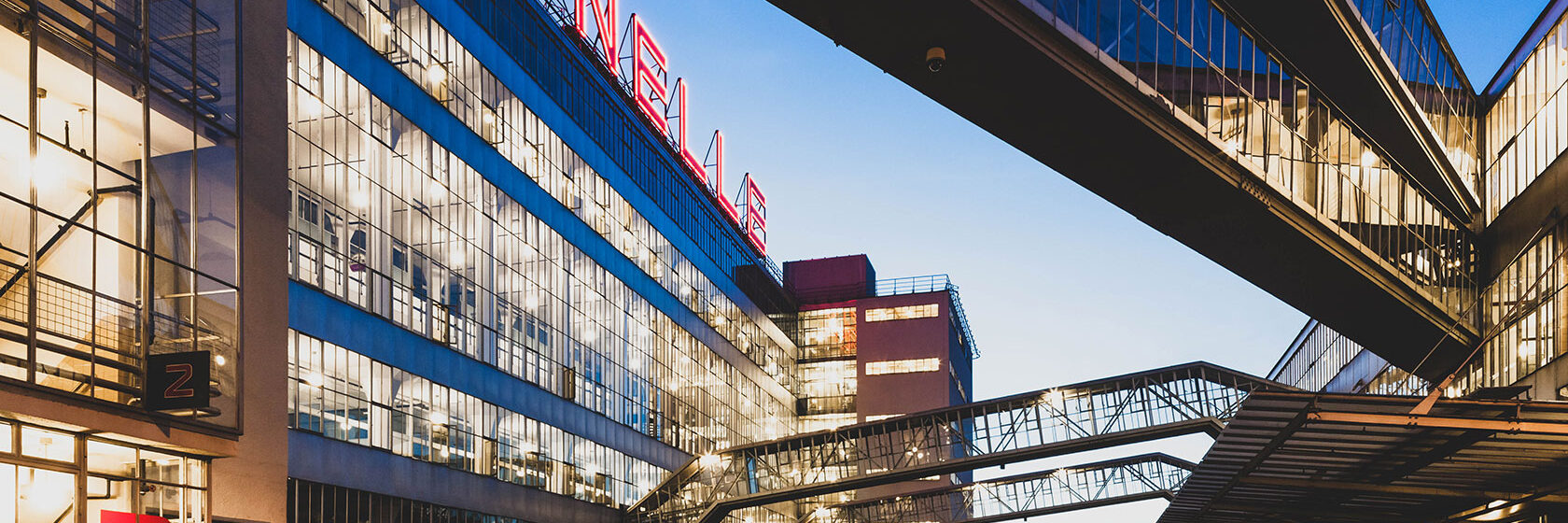 Modern byggnad med glasfasad och gångbroar under skymning, med neontecken "Van Nelle" högst upp.