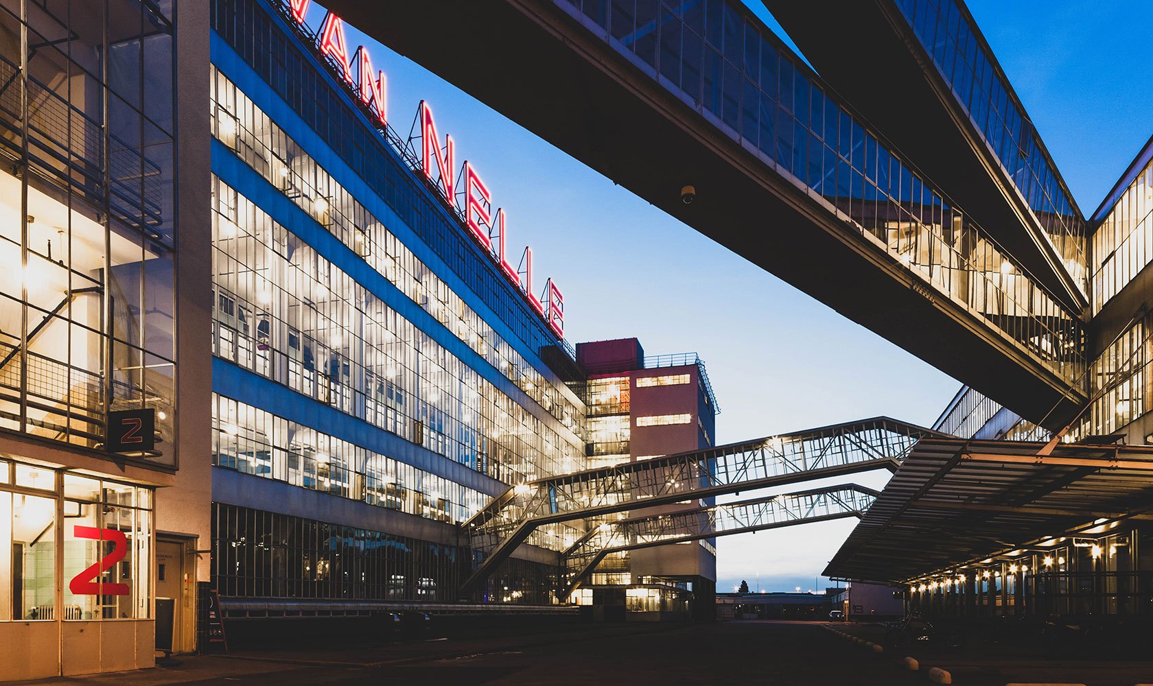 Modern byggnad med glasfasad och gångbroar under skymning, med neontecken "Van Nelle" högst upp.