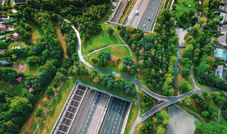 Flygbild av en motorväg med bilar och omgivande grönområde med gångvägar och träd.