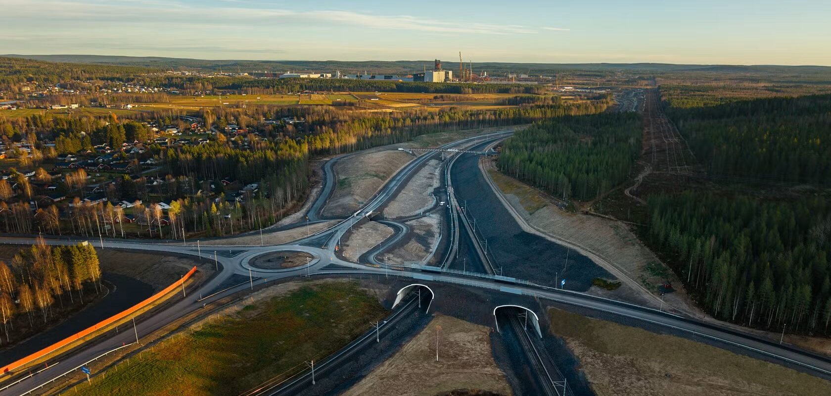 Flygfoto av en rondell och motorvägar nära en skog och en industribyggnad med blå himmel i bakgrunden.