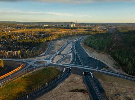 Flygfoto av en rondell och motorvägar nära en skog och en industribyggnad med blå himmel i bakgrunden.