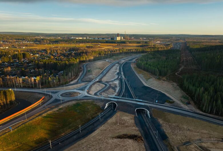 Flygfoto av en rondell och motorvägar nära en skog och en industribyggnad med blå himmel i bakgrunden.