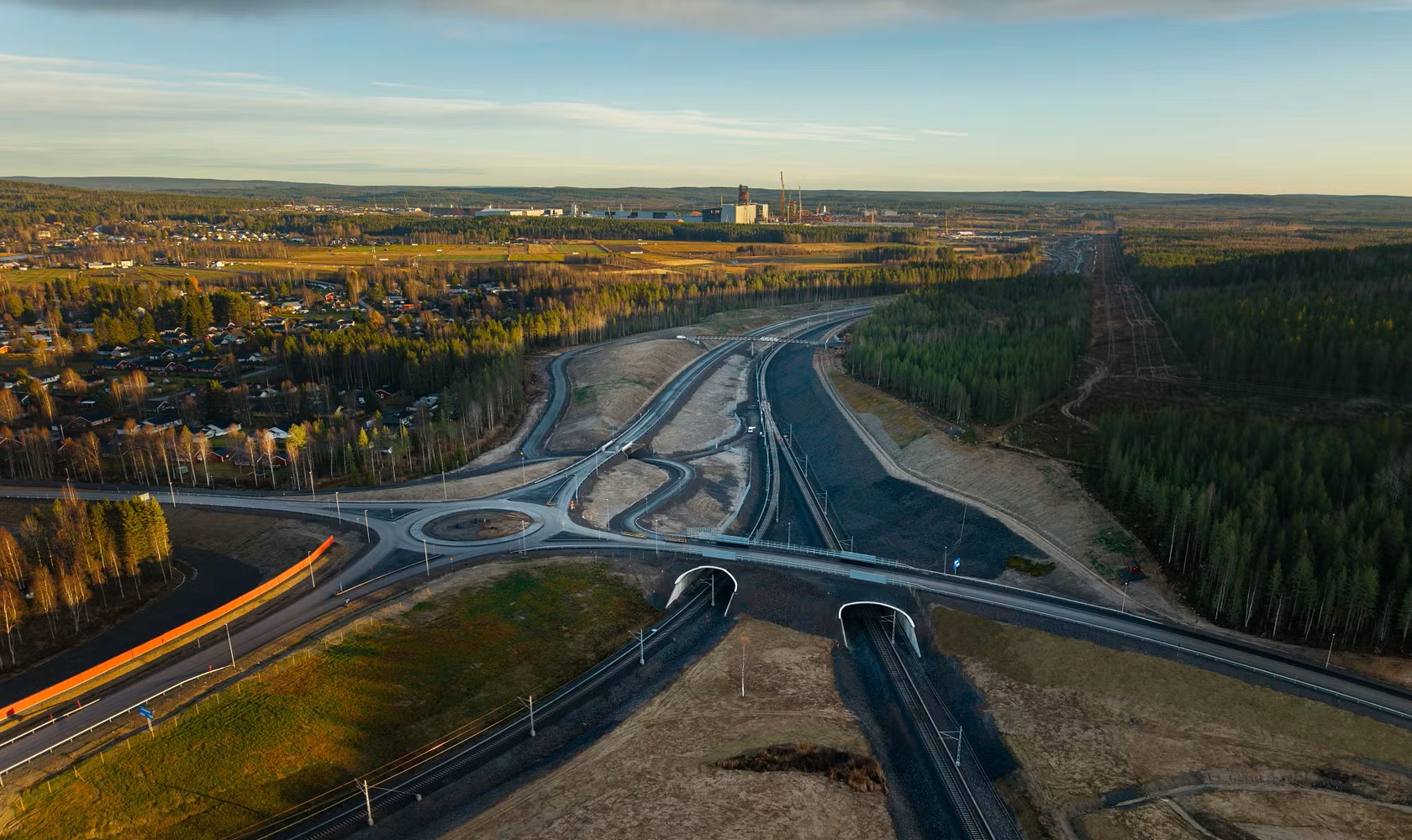 Flygfoto av en rondell och motorvägar nära en skog och en industribyggnad med blå himmel i bakgrunden.
