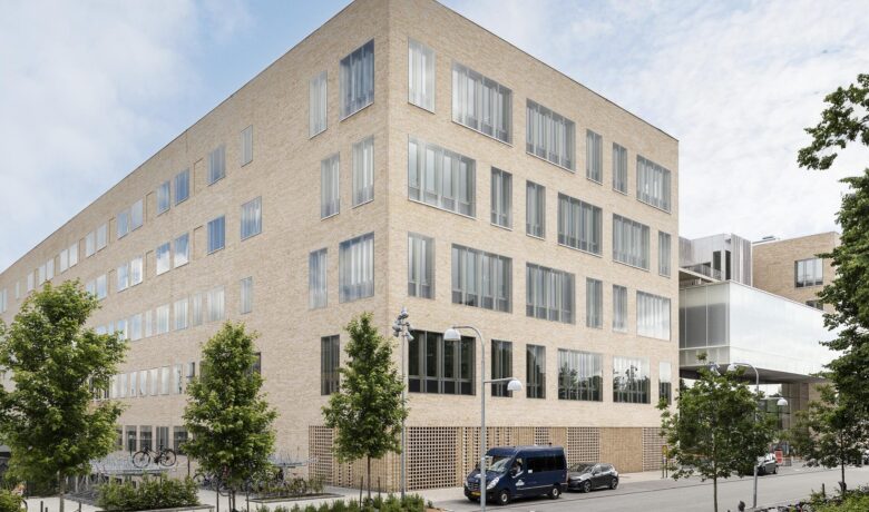 Modernt fyravåningshus med stora fönster, parkering och träd i närheten. Klar blå himmel i bakgrunden.