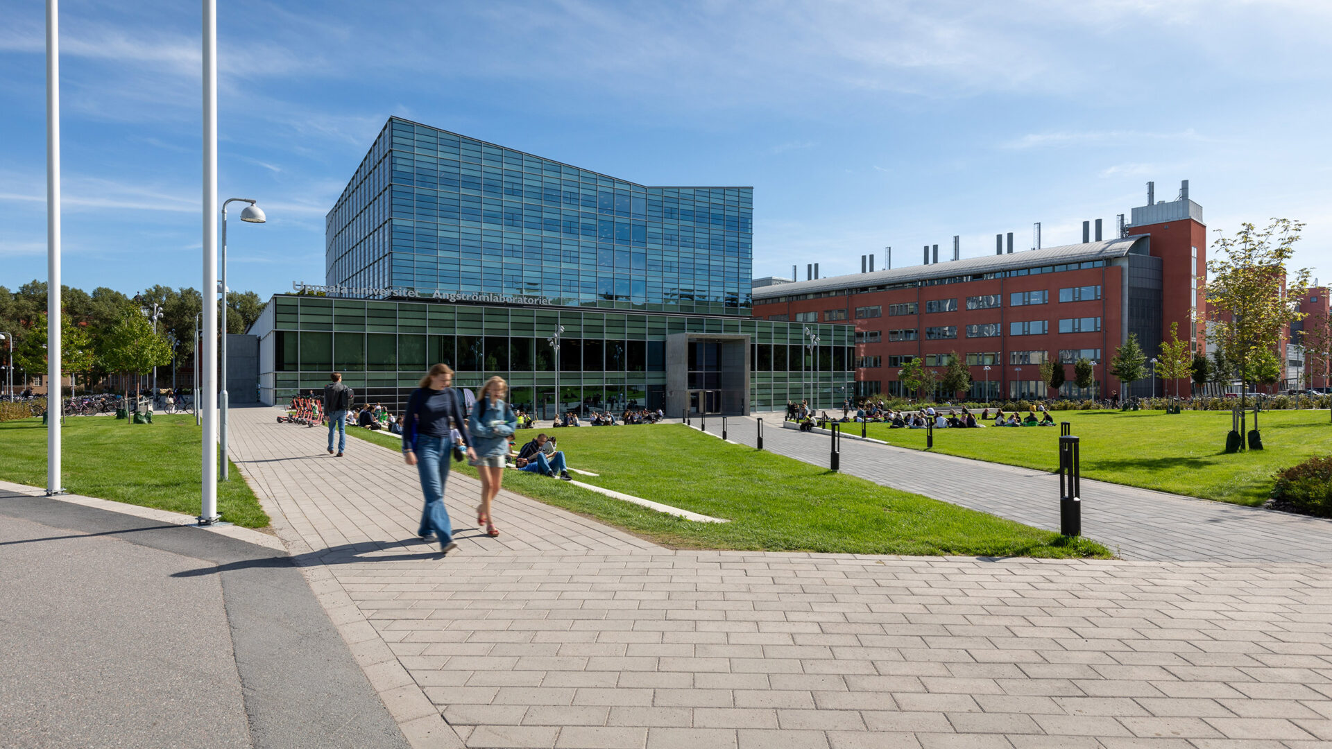 Modern universitetsbyggnad i glas vid en grön innergård, studenter promenerar och sitter på gräset under klar himmel.
