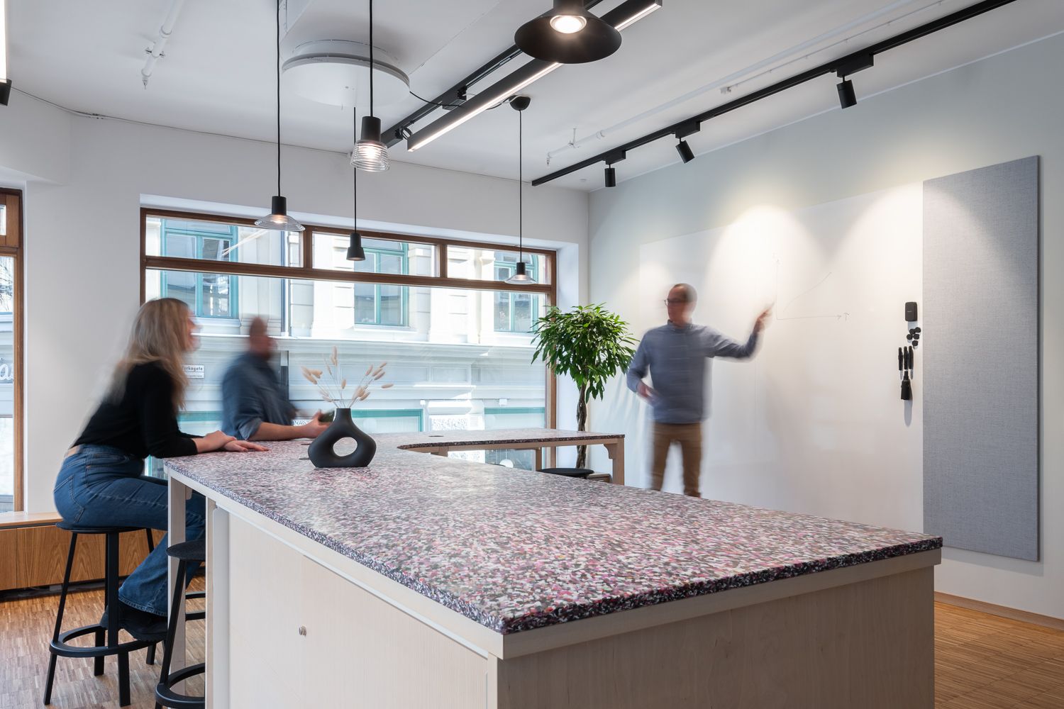 Modernt mötesrum med långt terrazzo-bord, barstolar, hängande lampor, stora fönster, växt och en person som skriver på en whiteboard.