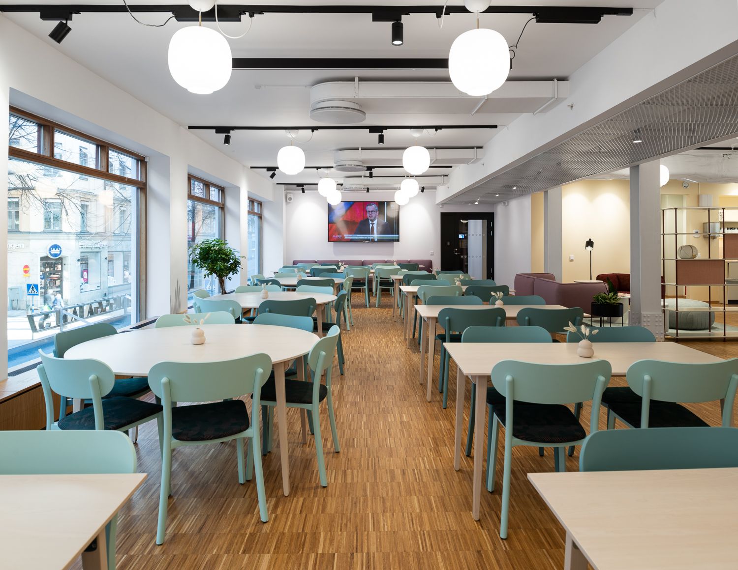 Ljust, modernt lunchrum med rader av bord och mintgröna stolar, stora fönster längs ena sidan och runda taklampor.