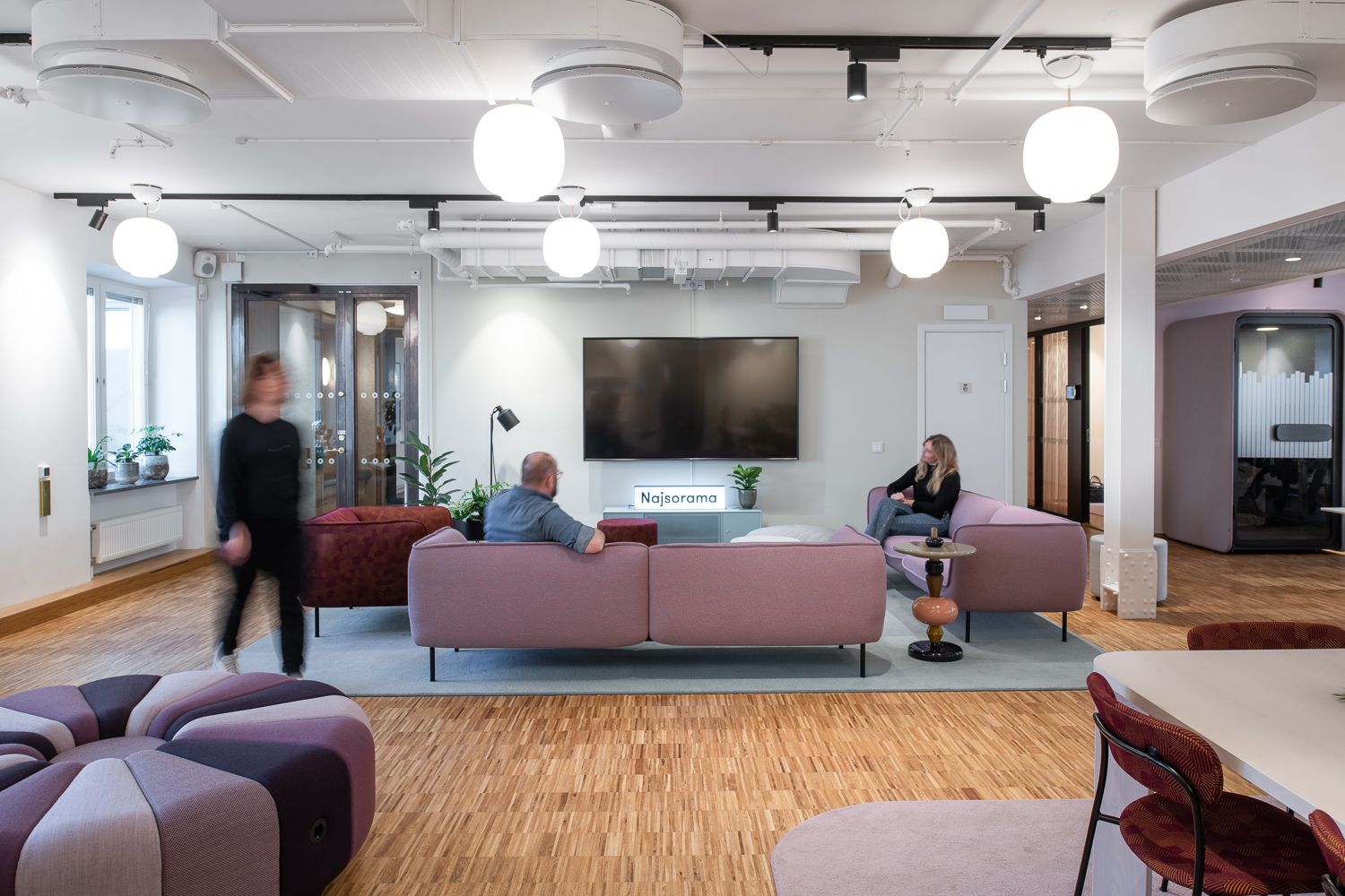 Öppet kontorslounge med rosa soffa, stor TV på väggen, runda taklampor och några personer.