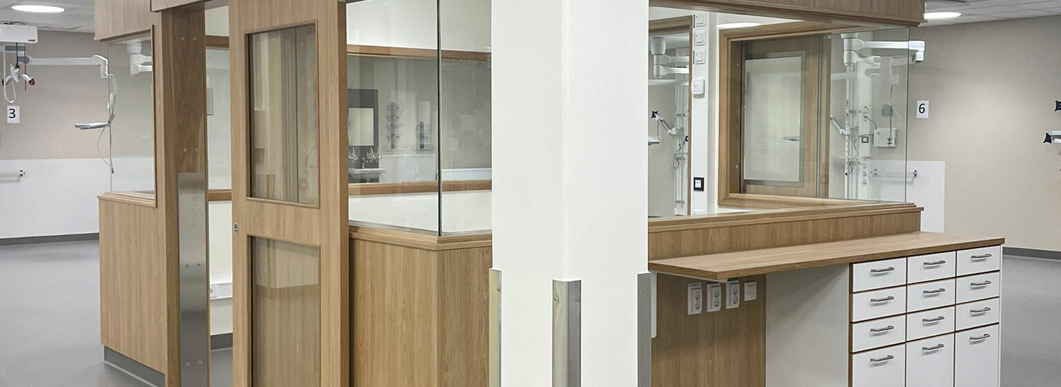 Modern sjukvårdsmiljö med träskåp och glaspaneler. Ljust golv och tak med vit panel.