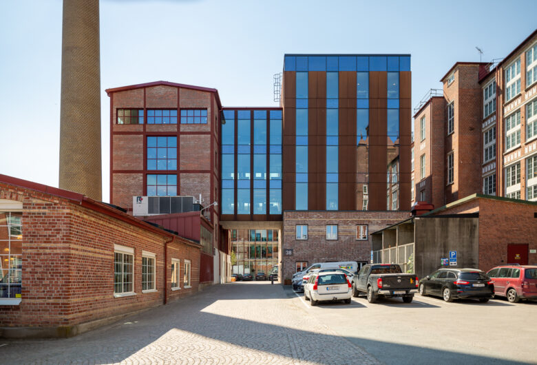 Gårdsbild med röda tegelbyggnader, hög skorsten, modern glas- och plåtfasad och parkerade bilar.