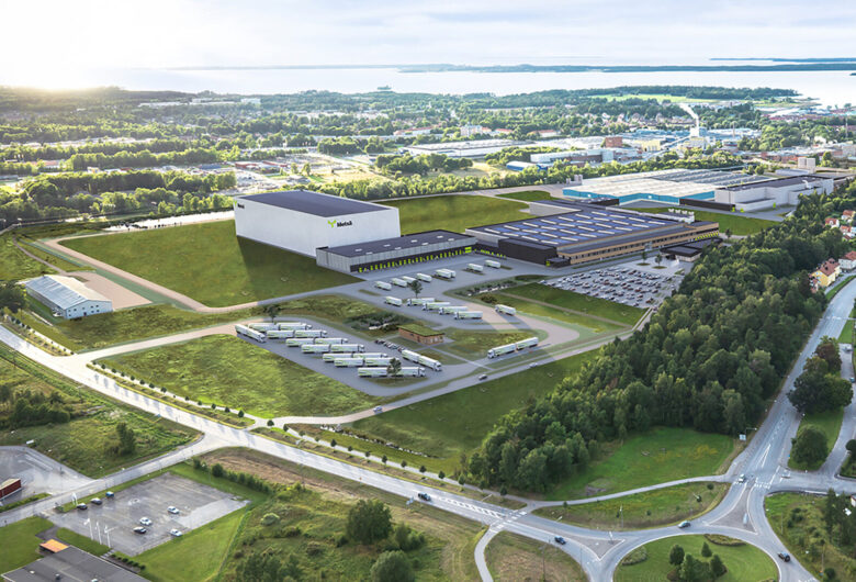 Flygfoto av en stor industribyggnad med lastbilar och parkering, omgiven av grönområden och småhus.