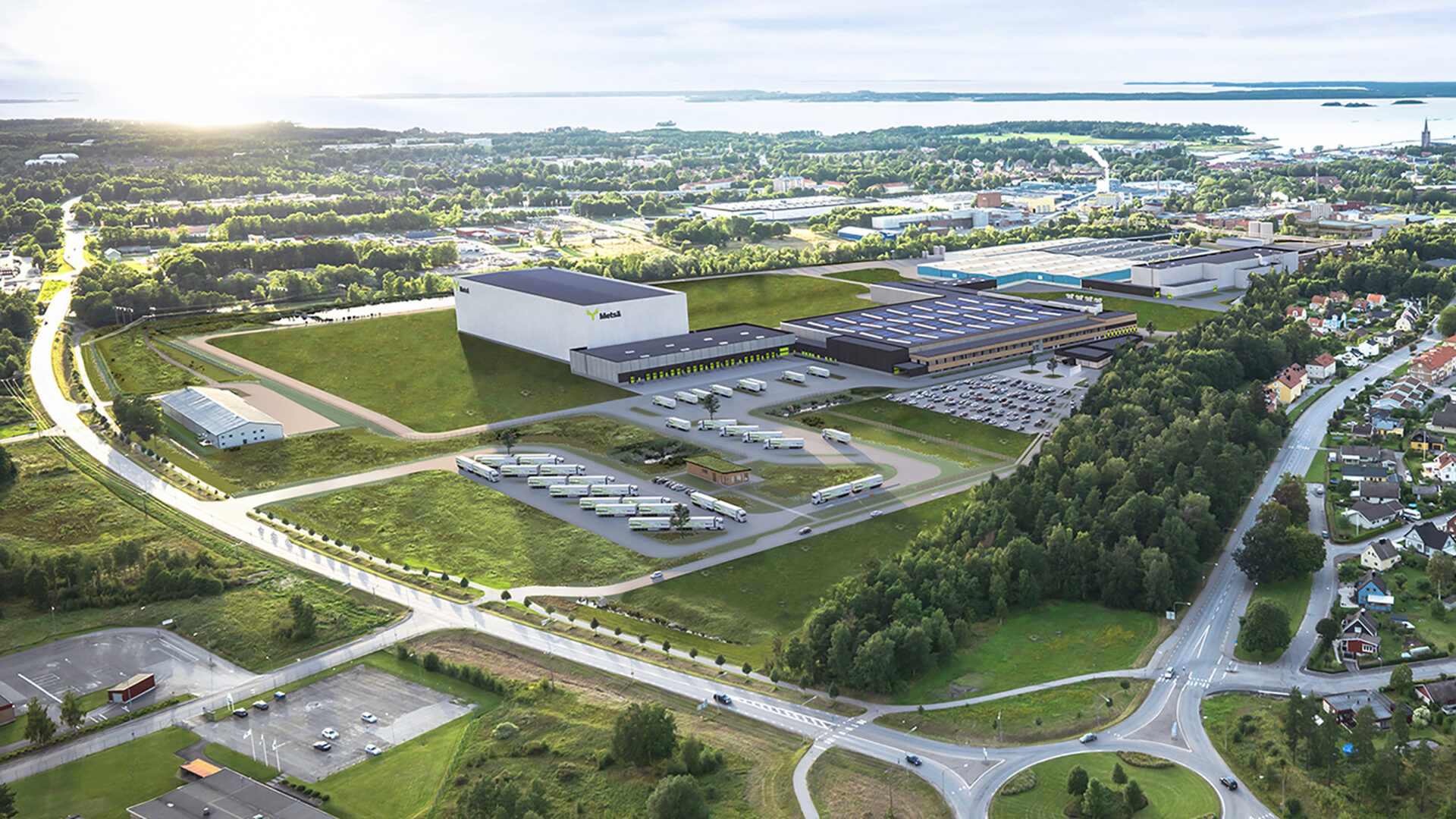 Flygfoto av en stor industribyggnad med lastbilar och parkering, omgiven av grönområden och småhus.