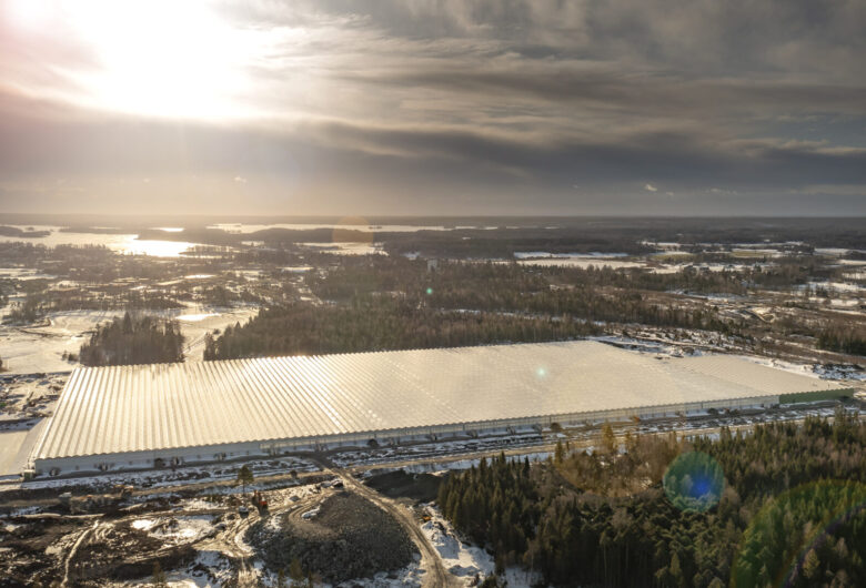 Flygfoto över stort reflekterande tak (växthus/industrihall) i snöklätt landskap under låg vintersol.