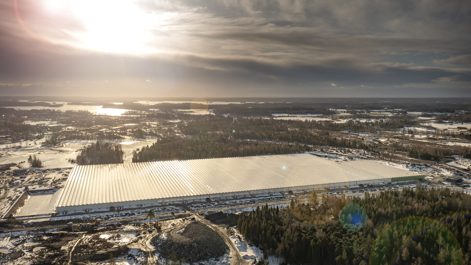 Flygfoto över stort reflekterande tak (växthus/industrihall) i snöklätt landskap under låg vintersol.