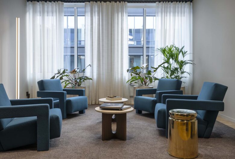 Modern sittgrupp med blå fåtöljer och två soffbord, inramad av stora fönster och gröna växter.