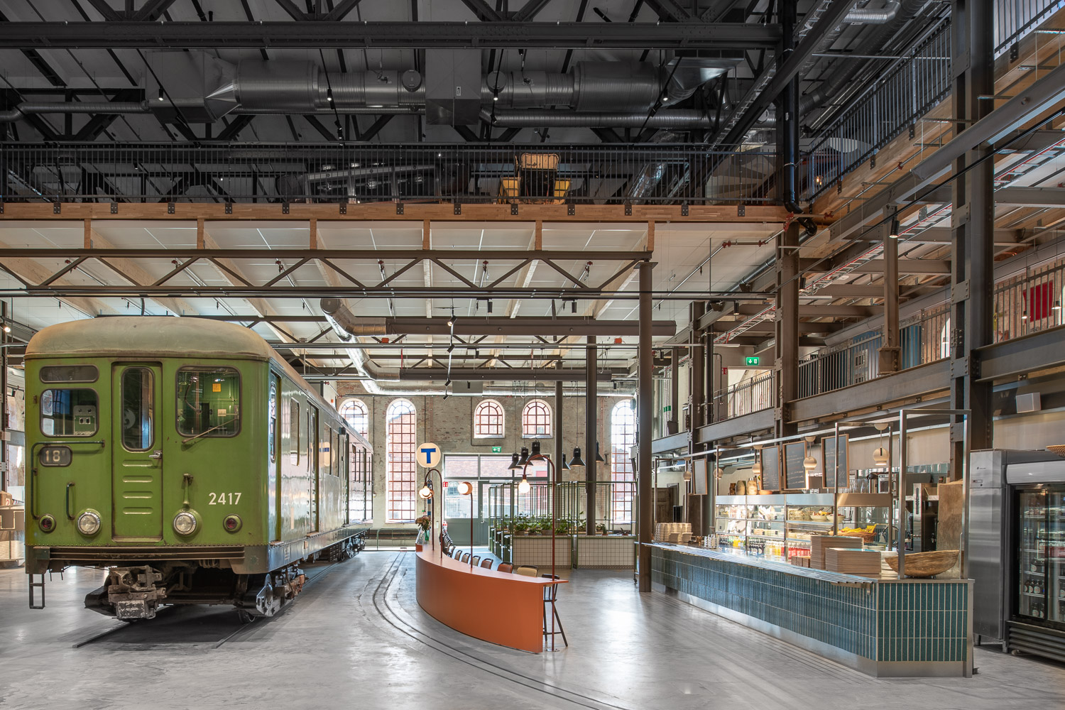 Interiör av ombyggd industribyggnad med grön spårvagnsvagn på räls, böjd orange disk, café och mezzanin med stora bågade fönster.