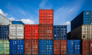 Blå, röda och grå lastcontainrar staplade på varandra med blå himmel i bakgrunden.