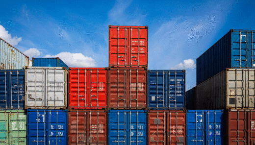 Blå, röda och grå lastcontainrar staplade på varandra med blå himmel i bakgrunden.