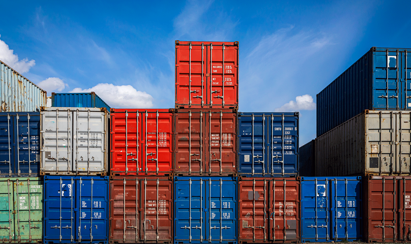 Blå, röda och grå lastcontainrar staplade på varandra med blå himmel i bakgrunden.