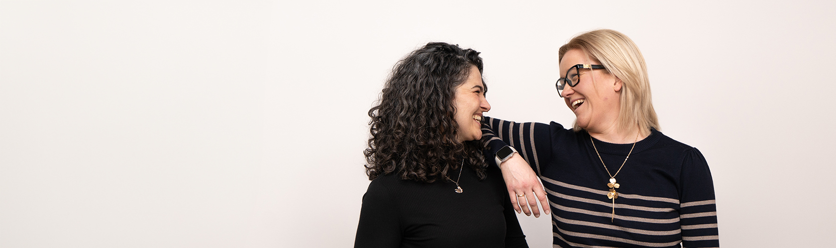 Två kvinnor stående nära varandra, båda bär halsband och ser glad ut.