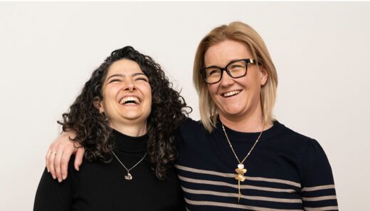 Två kvinnor stående nära varandra, båda bär halsband och ser glada ut.