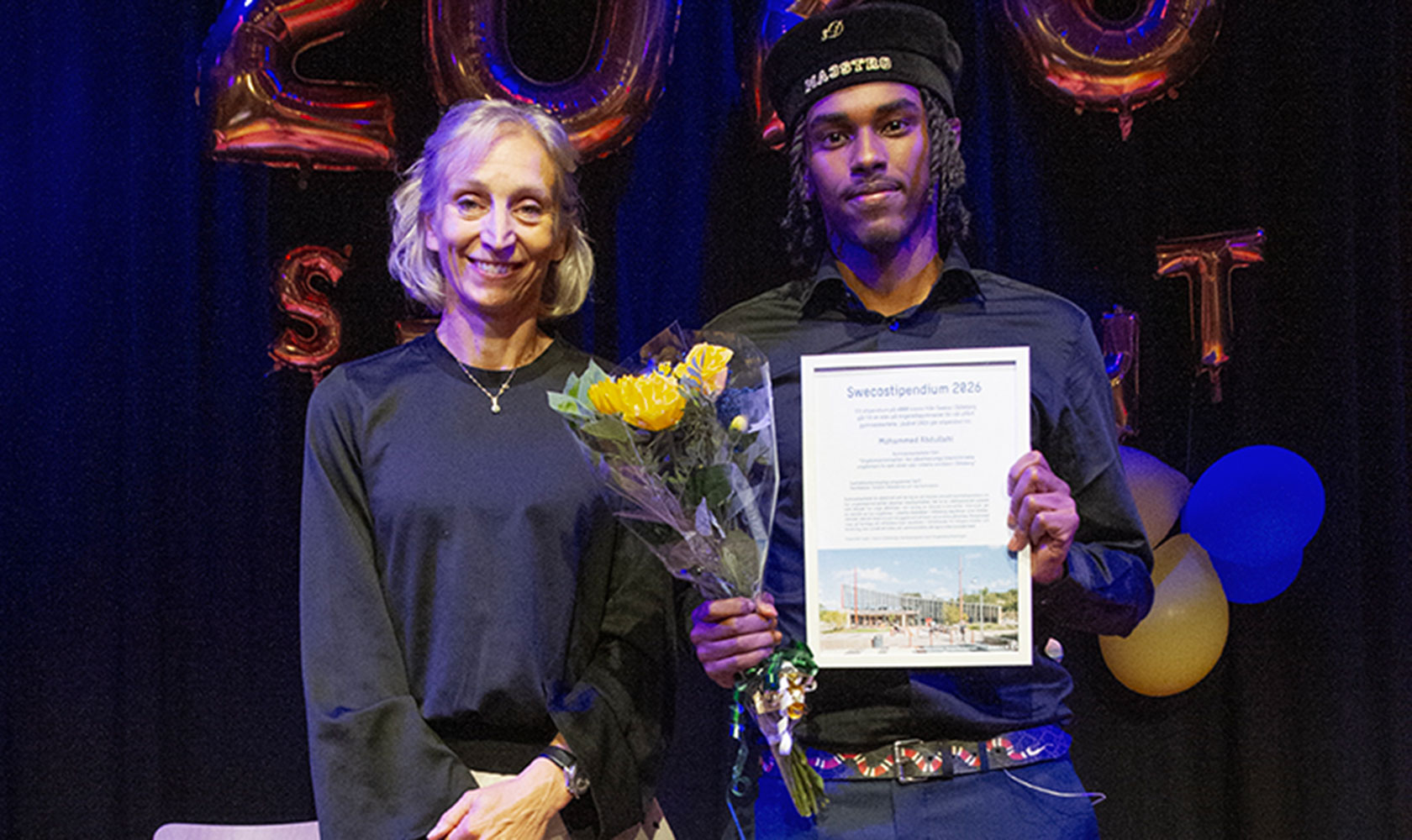 Två personer på prisutdelning med blommor och diplom, festballonger i bakgrunden