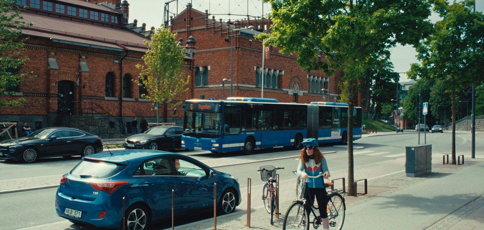 Cyklist på trottoar vid blå buss och tegelbyggnad i stadsmiljö med träd