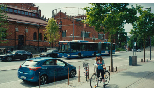 Cyklist på trottoar vid blå buss och tegelbyggnad i stadsmiljö med träd