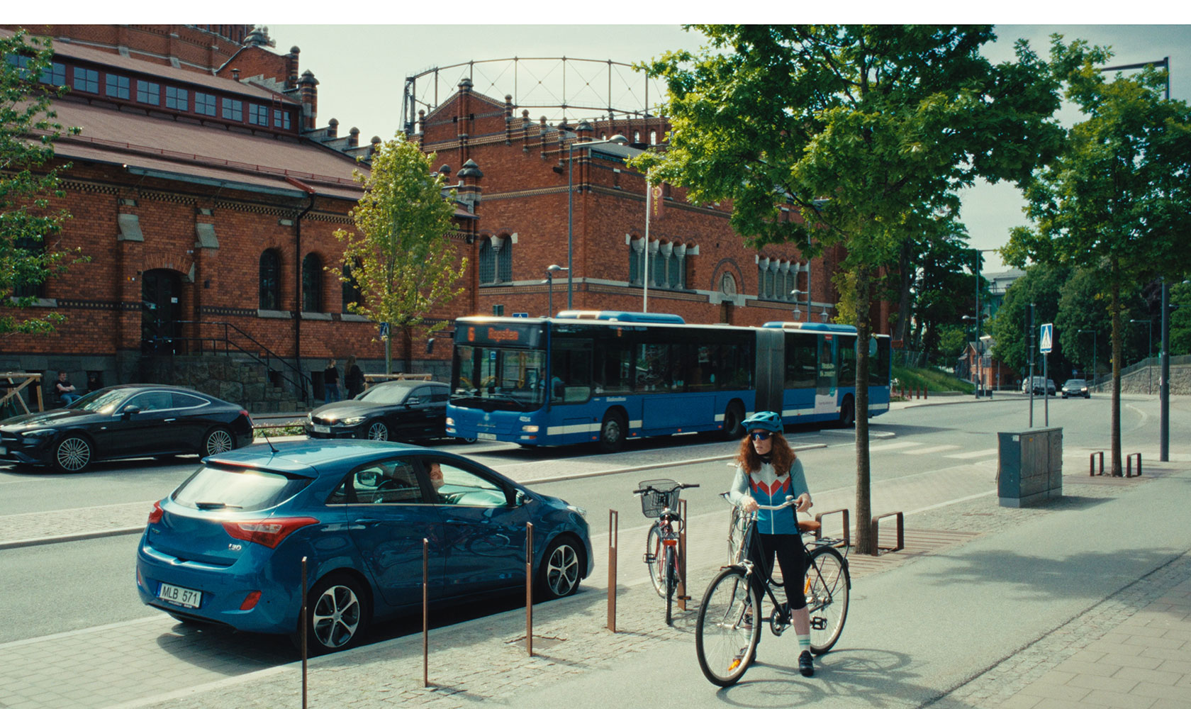 Cyklist på trottoar vid blå buss och tegelbyggnad i stadsmiljö med träd