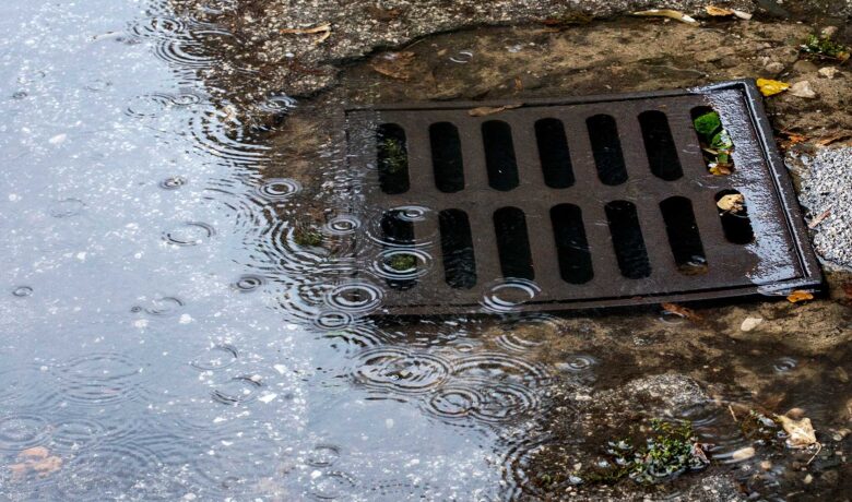 Regnvatten rinner ner i gatubrunn på våt asfalt med ringar i vattenpöl